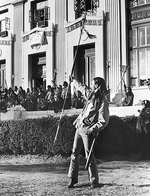 Native American man holds up a spear as others gather in front of a government building.
