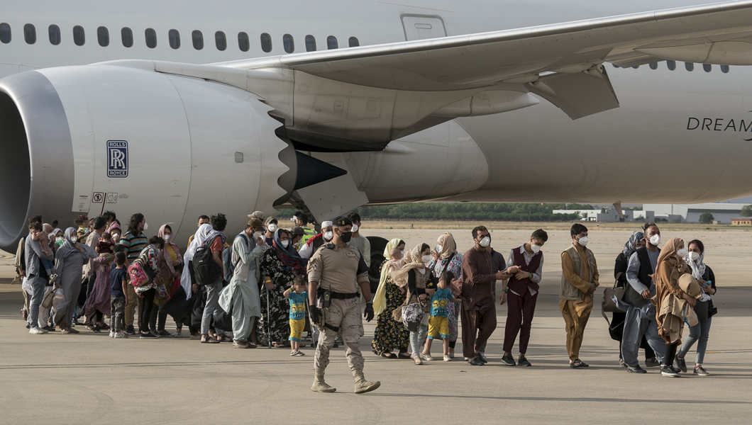A line of masked passengers waiting to board a plane, overseen by an armed guard in uniform.