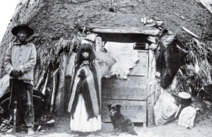 Man and woman of the Havasupai tribe standing in front of a small hut.