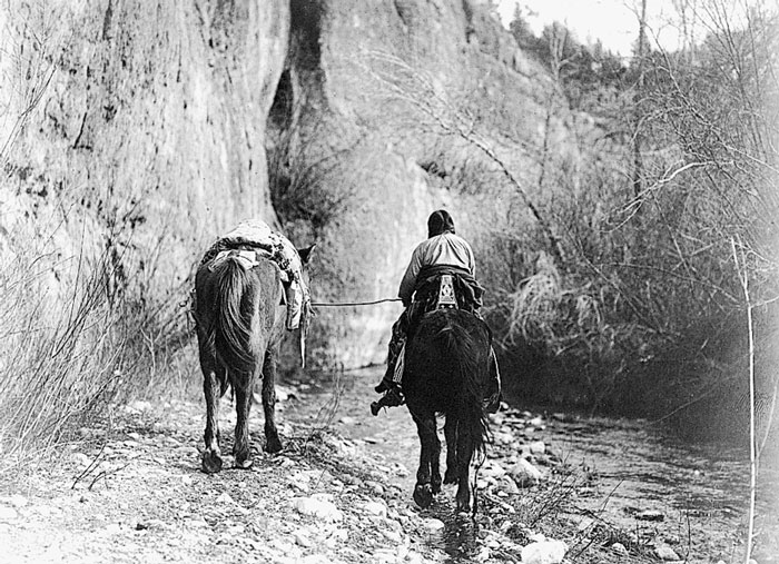 Native America rides a horse through a valley while guiding another horse along side of him.