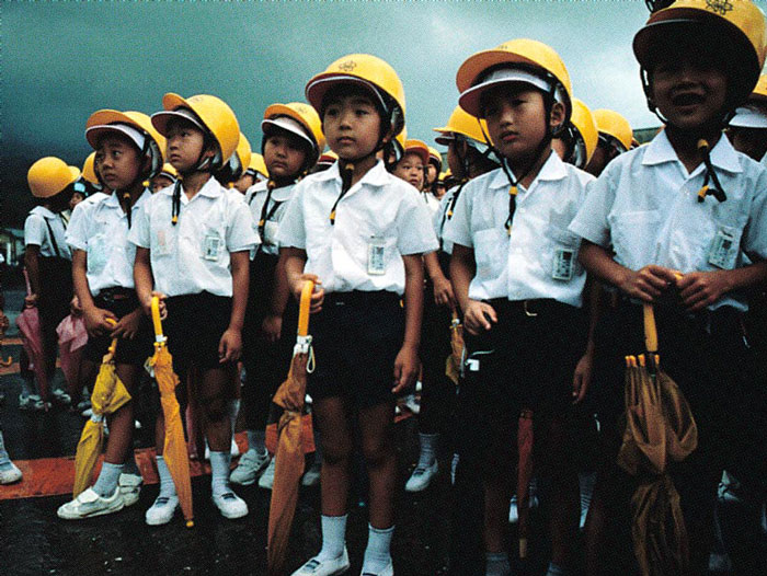 Japanese students wearing protective helmets and carrying umbrellas.