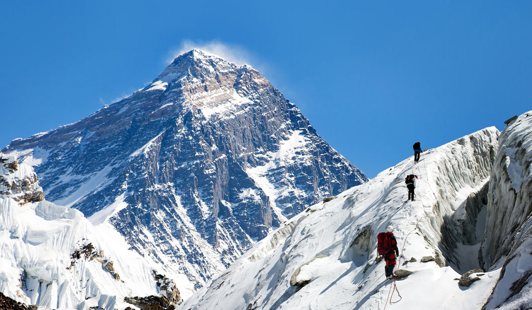 Three climbers hike up a steep snowy slope with a very tall mountain in the background.
