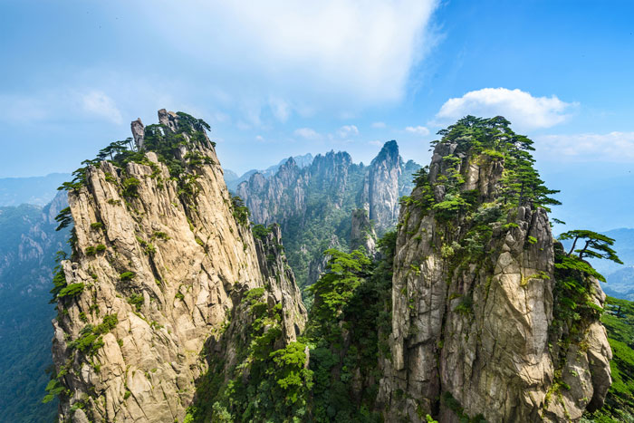 Two narrow mountains next to each other with trees growing on them. Other mountains are seen behind them. 