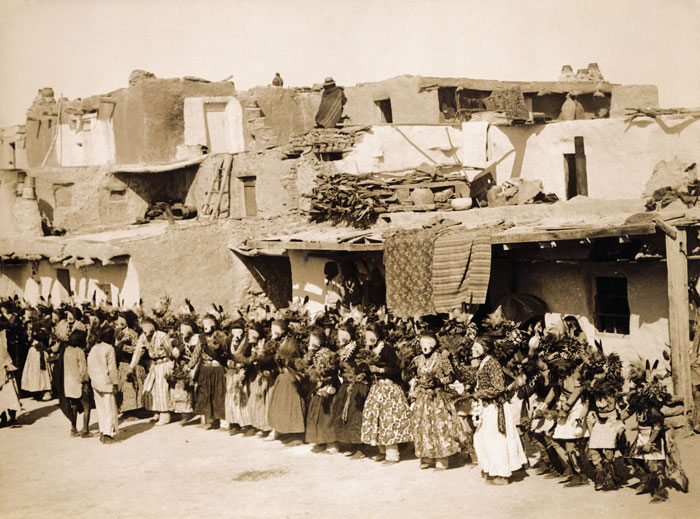 A large crowd of people wearing masks stand in front of a pueblo.