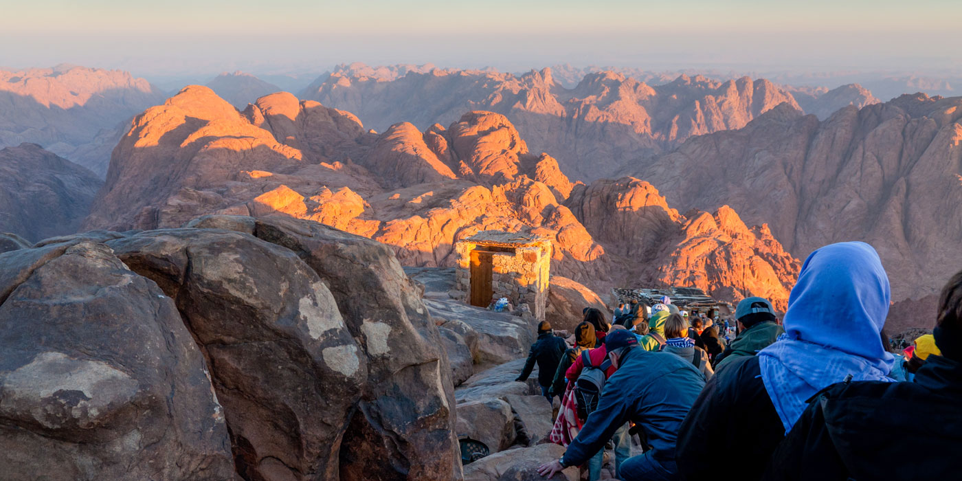 People walk down a path on top of a mountain. The view looks out at other mountains in the distance. 