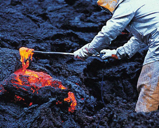 Scientist wearing protective clothing uses a long scoop to colect lava.