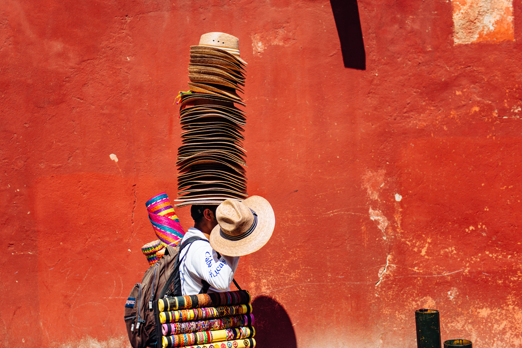 Street vendor walking down the street wearing a tall pile of hats on his head. 