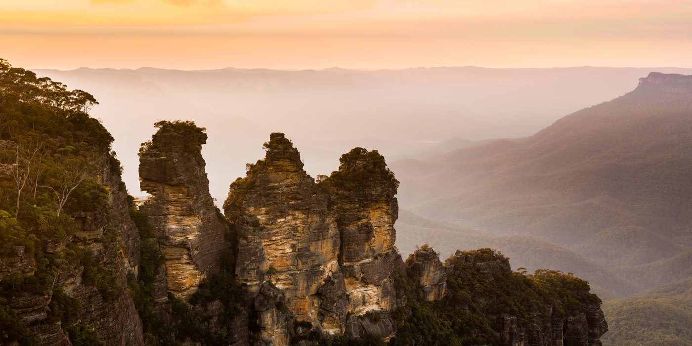 Three small mountain peaks stick up next to each other with the view behind them of a valley and other mountains. 
