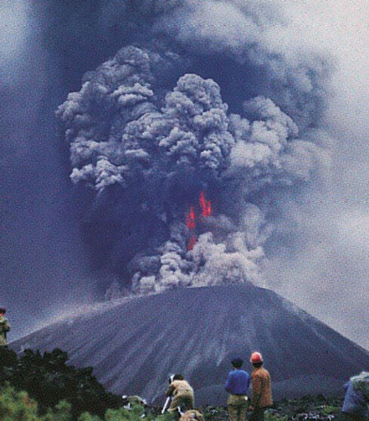 Three scientists stand in the distance observing an erupting volcano.