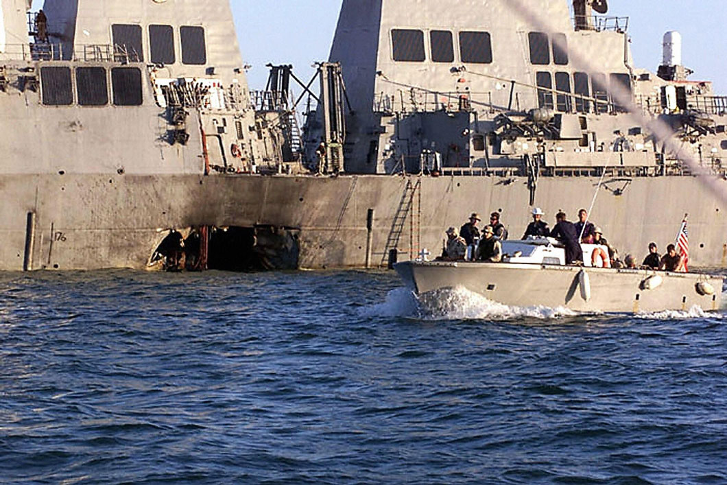 A small motorboat with uniformed and civilian personnel near a large vessel with a large hole in its side.