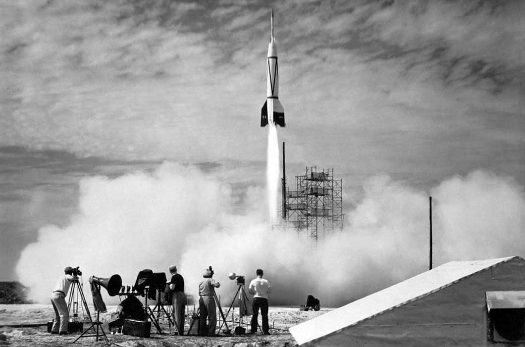 Missle being launched into the air as people stand around it to observe. 