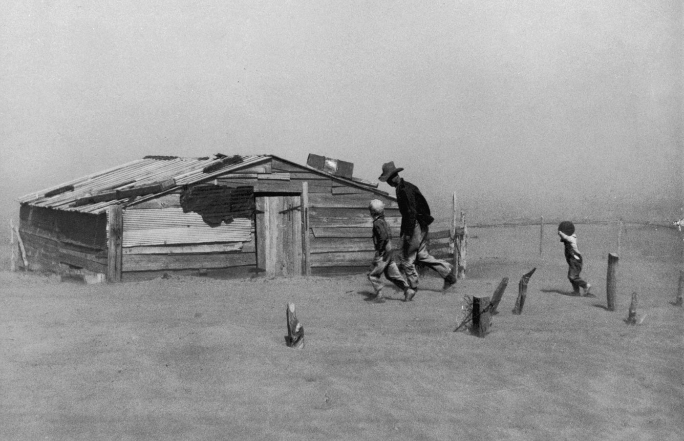 Man and two children walk past a rundown shack as dust blows in the air.