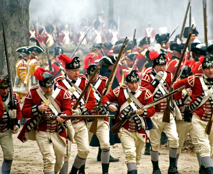 Men dressed as British soldiers with muskets advance with their guns pointed. 