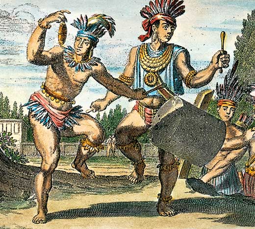 Two Native Americans wearing feathers beat a drum.