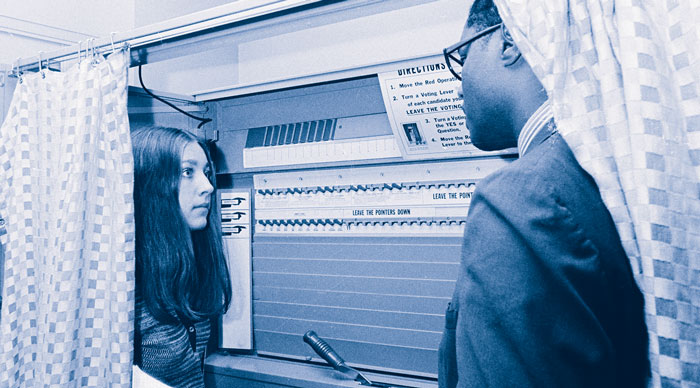 Photo of a young woman and a man standing facing one another in a voting booth with the curtains open.