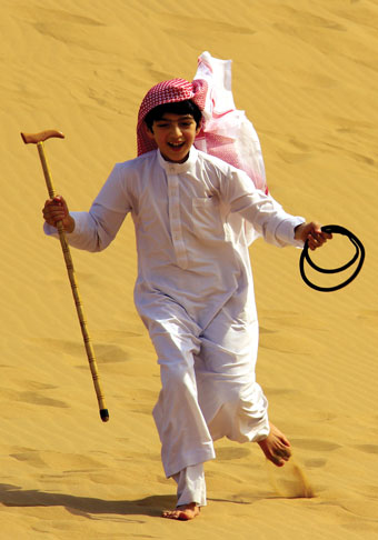 Yong boy wearing white longsleeved clothing and a hat walks in the desert carrying a cane.